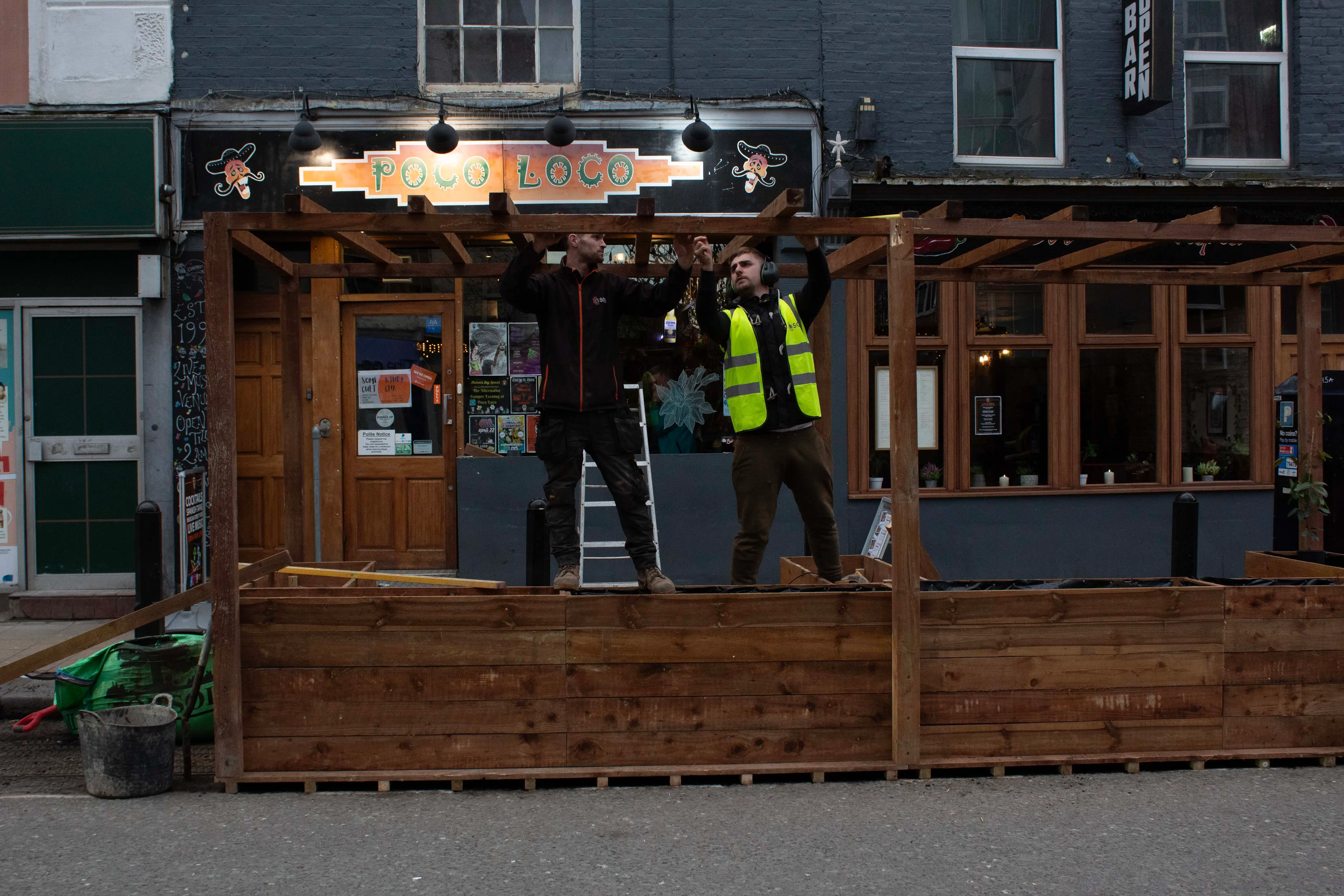 Two workers in safety gear stand outside a wooden structure with lights, preparing for a job. Cityscape backdrop.