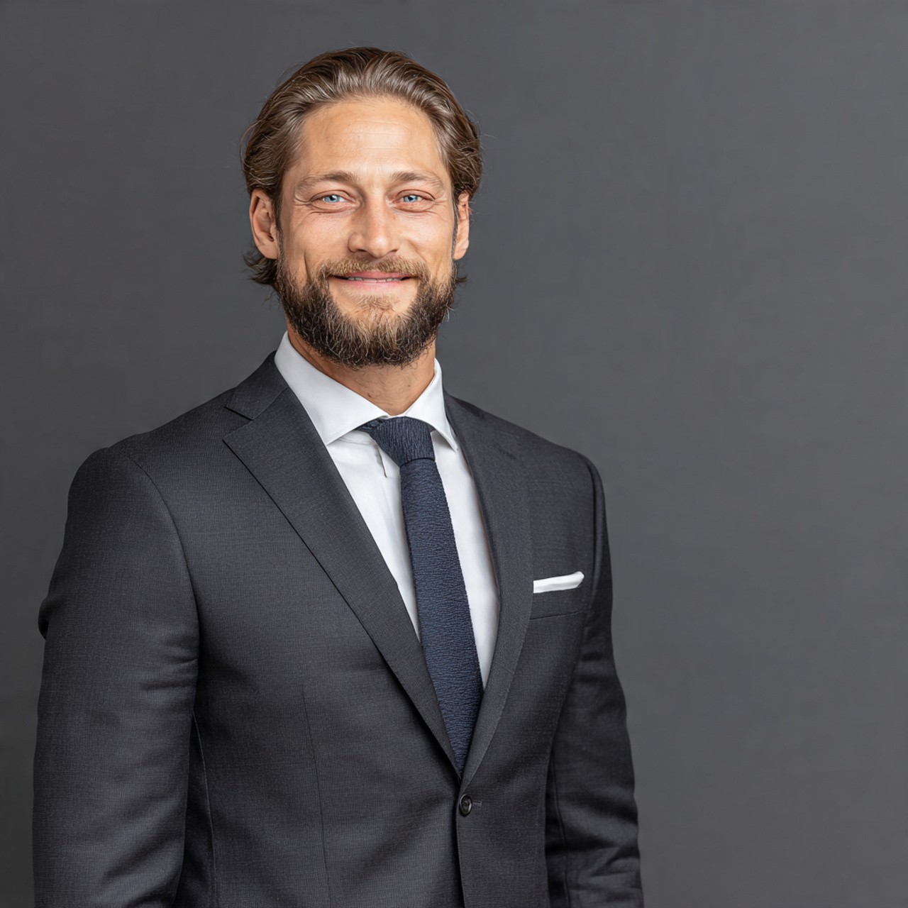 Smiling man with a beard in a dark suit and tie, standing against a gray background. He appears confident and professional.