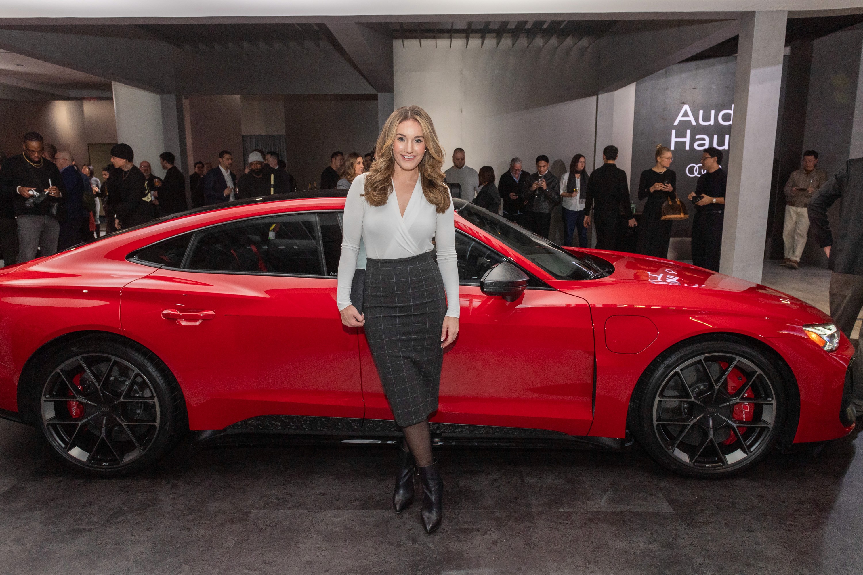 A woman stands smiling in front of a sleek red Audi car at an indoor showroom event. People mill about in the background, creating an energetic atmosphere.