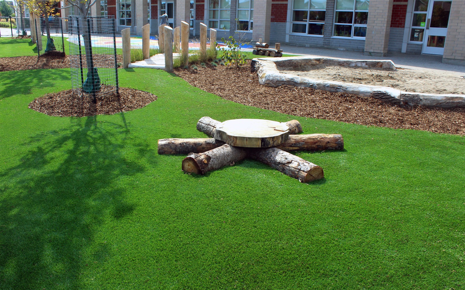 A lush green playground featuring a faux wooden fire pit made of tree stumps and stones, creating a cozy outdoor make-believe atmosphere.