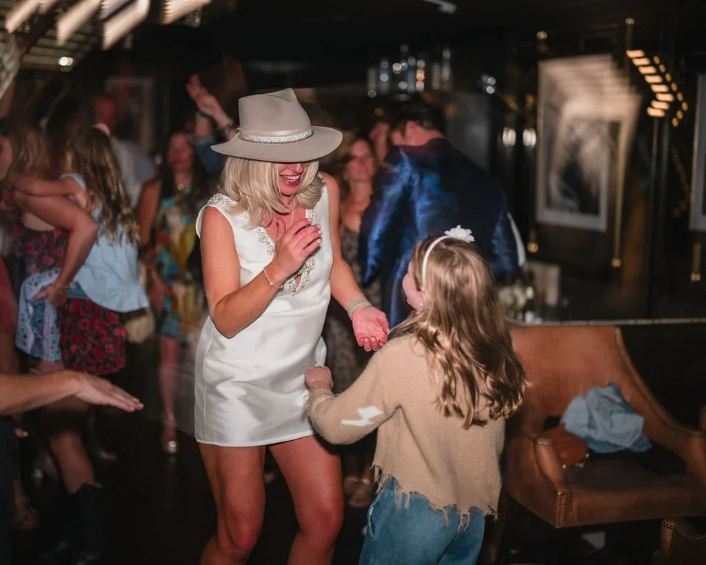 The bride in a short white dress and wide-brimmed hat dances with a young guest.