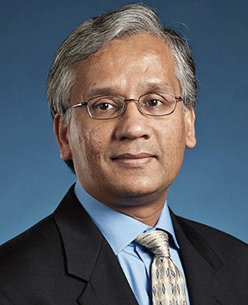 A professional headshot of a man with glasses, wearing a suit and tie, in front of a blue background.