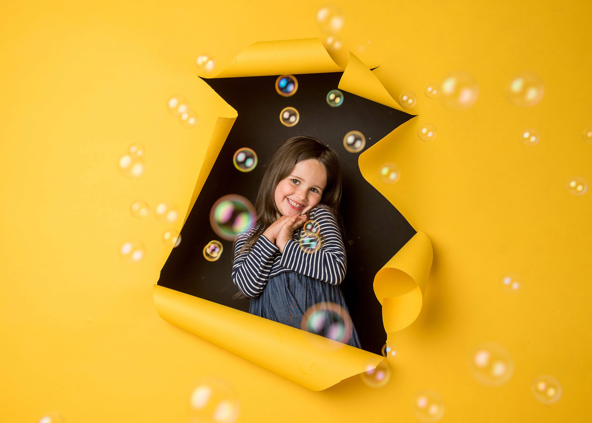 Kreative moderne Kitafotografie Gevelsberg: Fröhliches Mädchen blickt durch rissiges gelbes Papier, umgeben von schwebenden bunten Seifenblasen bei PK photography 4 Kids.