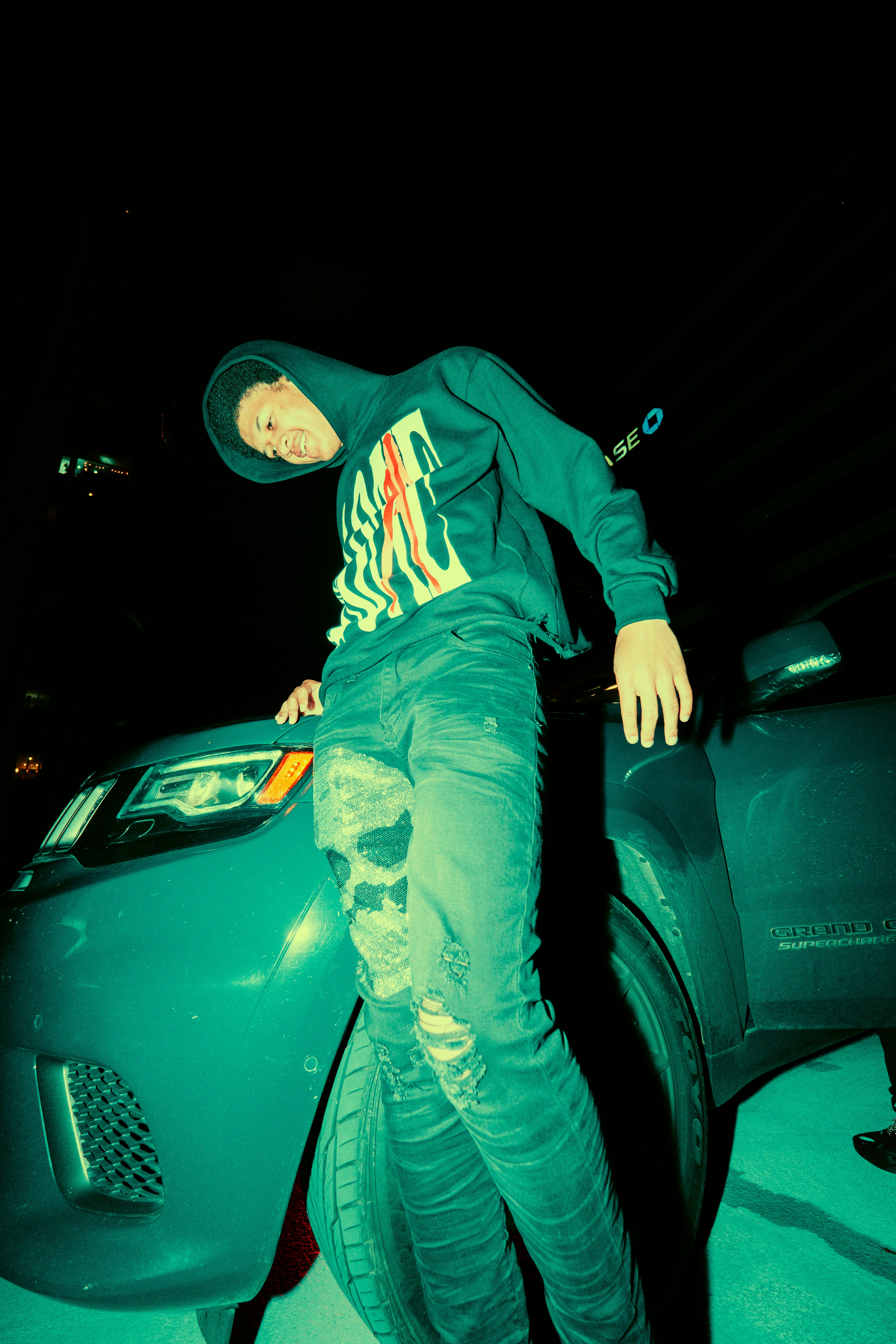 man in black jacket and green pants wearing black hat sitting on black car