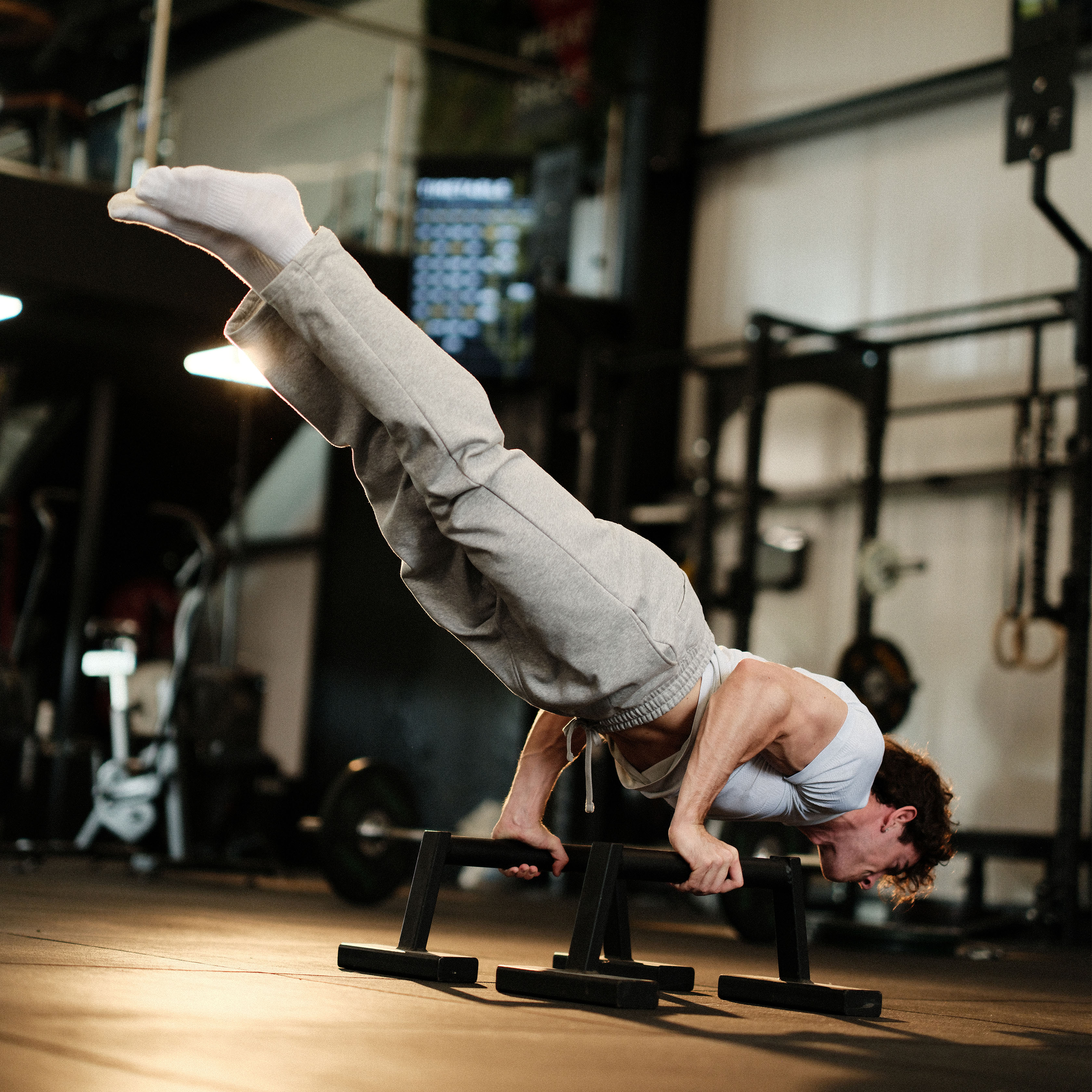 A muscular man smiling while performing a hanging leg raise on gymnastic rings in a bright fitness studio.