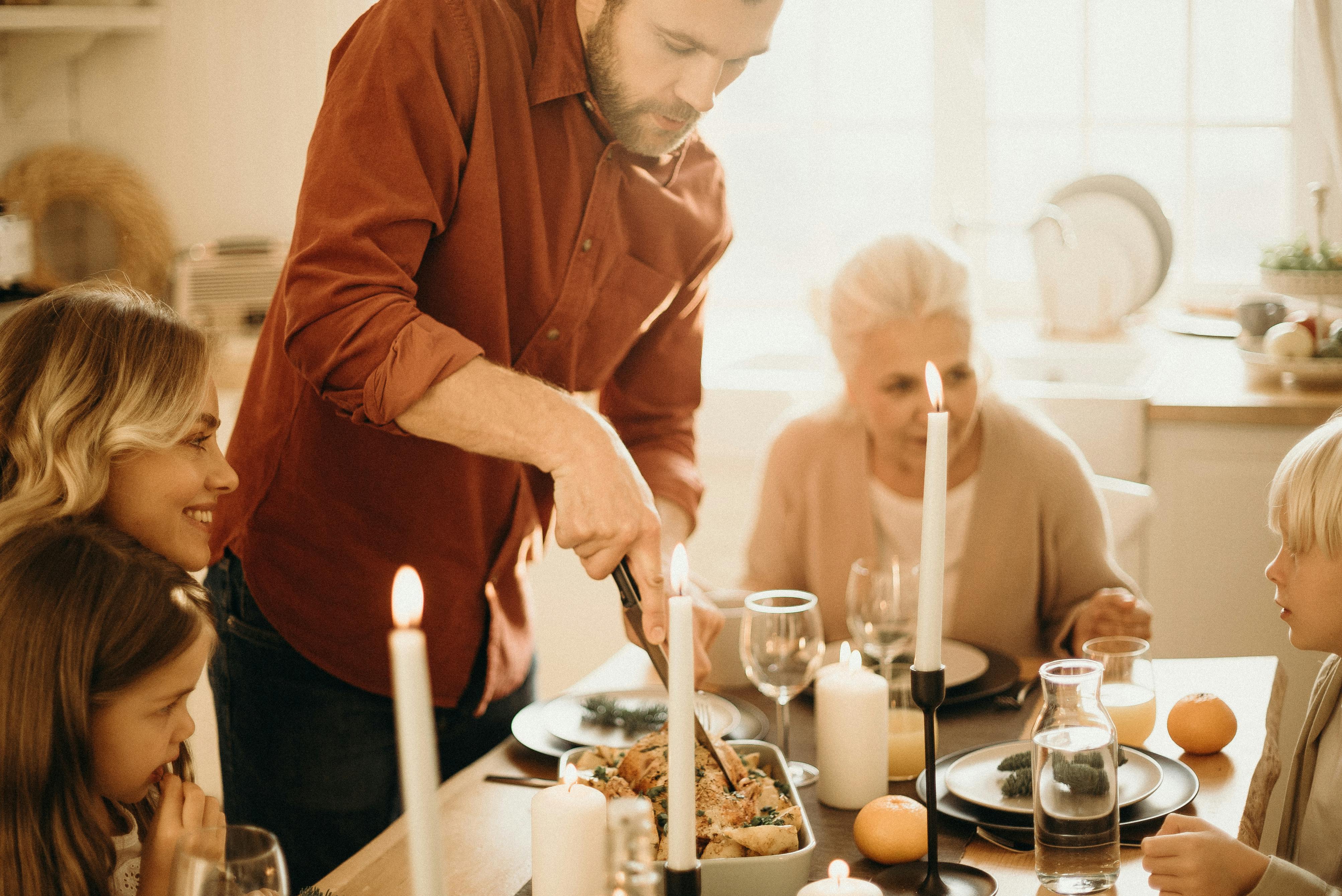 Family gathered around a candlelit dinner table as a man carves a roasted chicken.