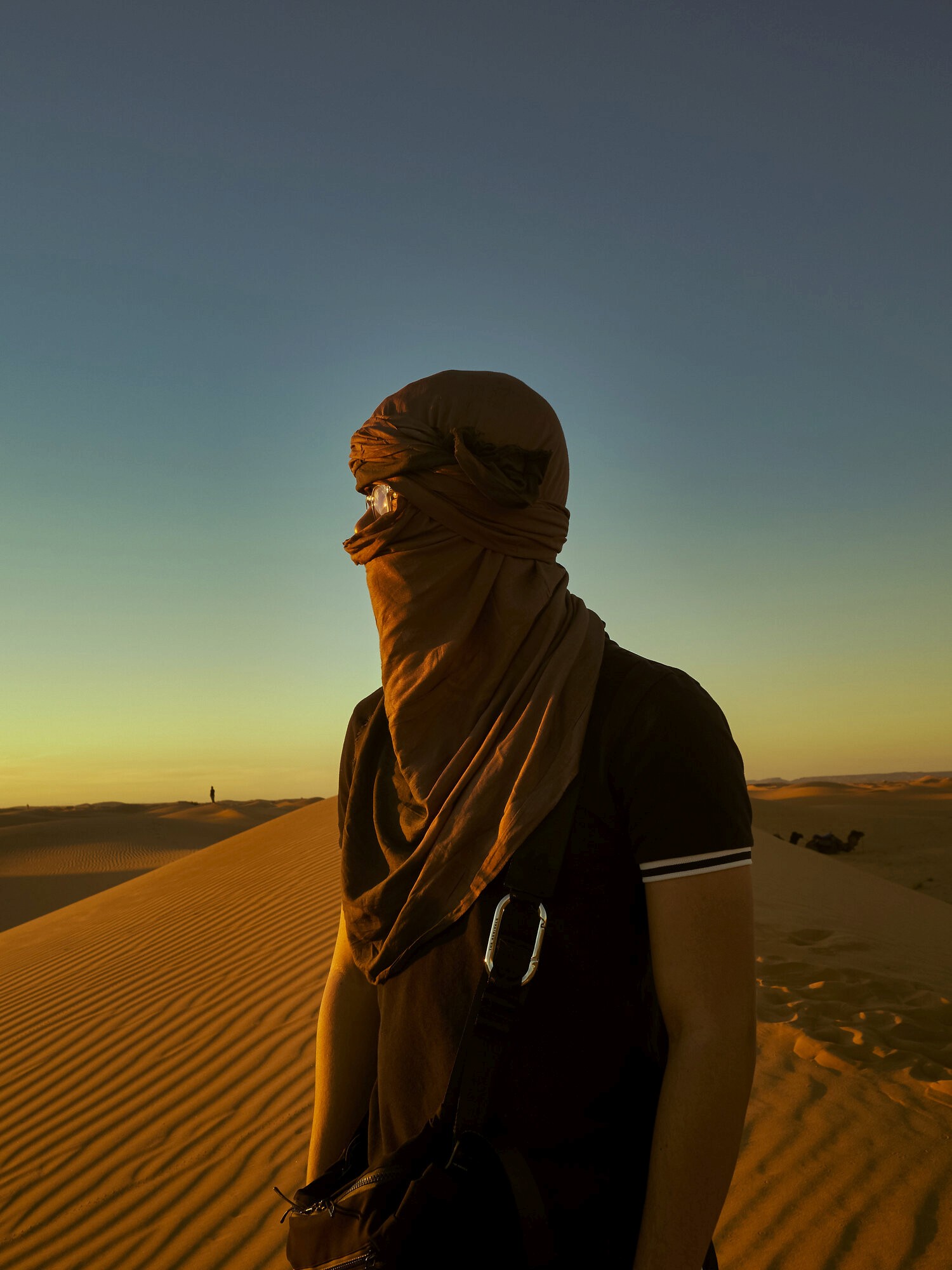 Person in a brown scarf on a sand dune at sunset in Morocco’s desert, casting long shadows in the golden light.