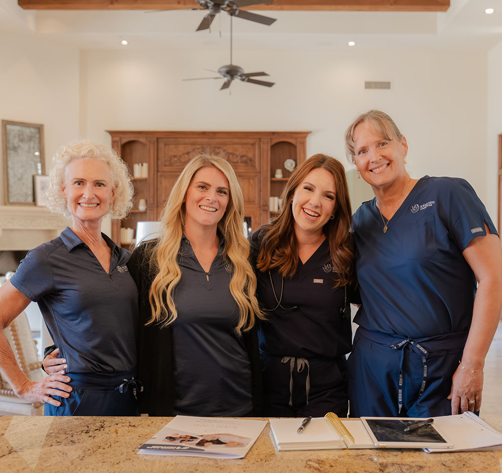 Four women in dark blue medical scrubs smiling and standing behind a counter with papers and pens on it.