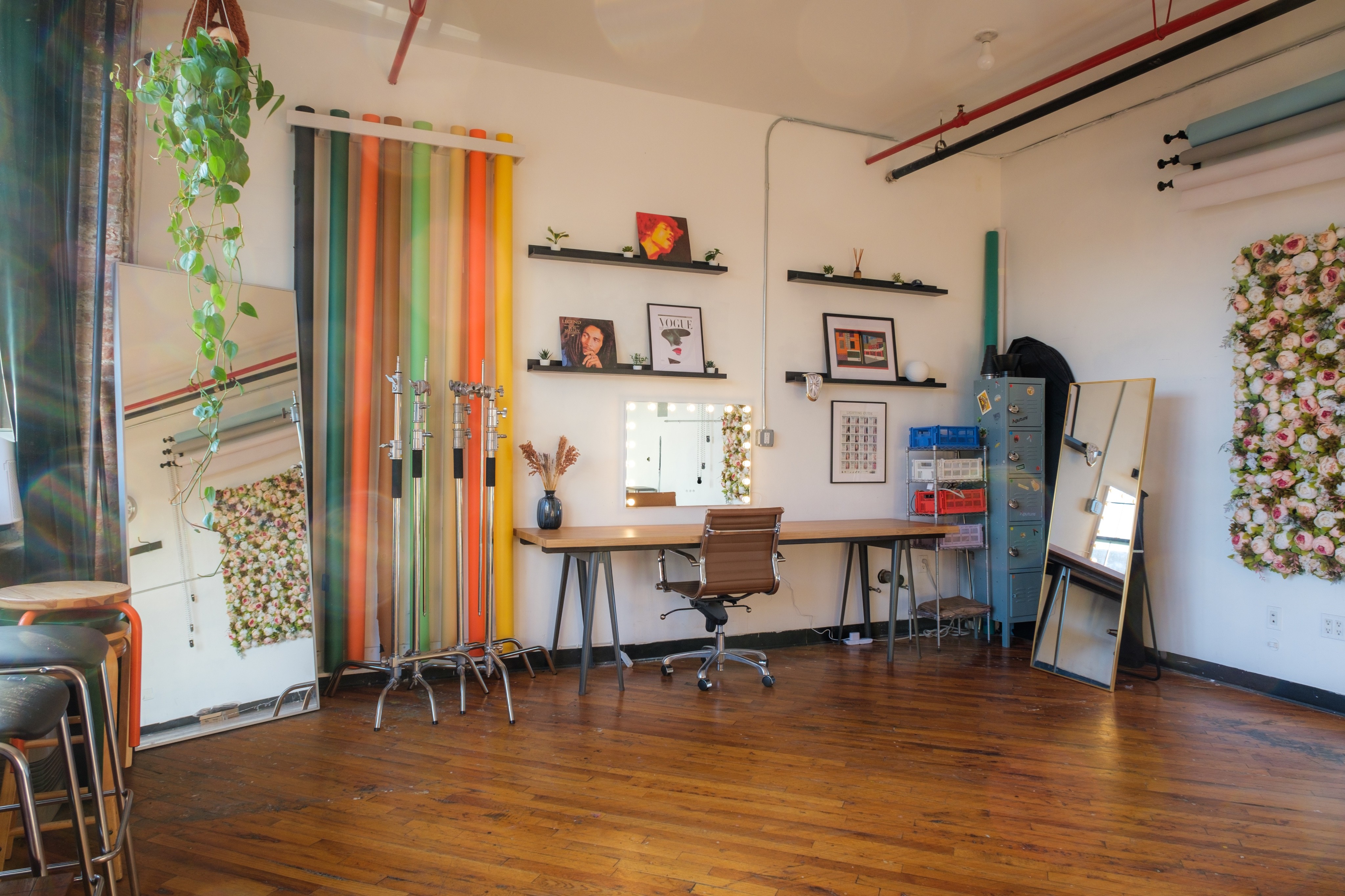 A workspace in a daylight photography studio, featuring a long wooden desk, an ergonomic chair, and lighting equipment set against colorful backdrops. The area includes shelves with decor, a floral-framed mirror with bulbs, and a storage unit, combining functionality with creative flair.