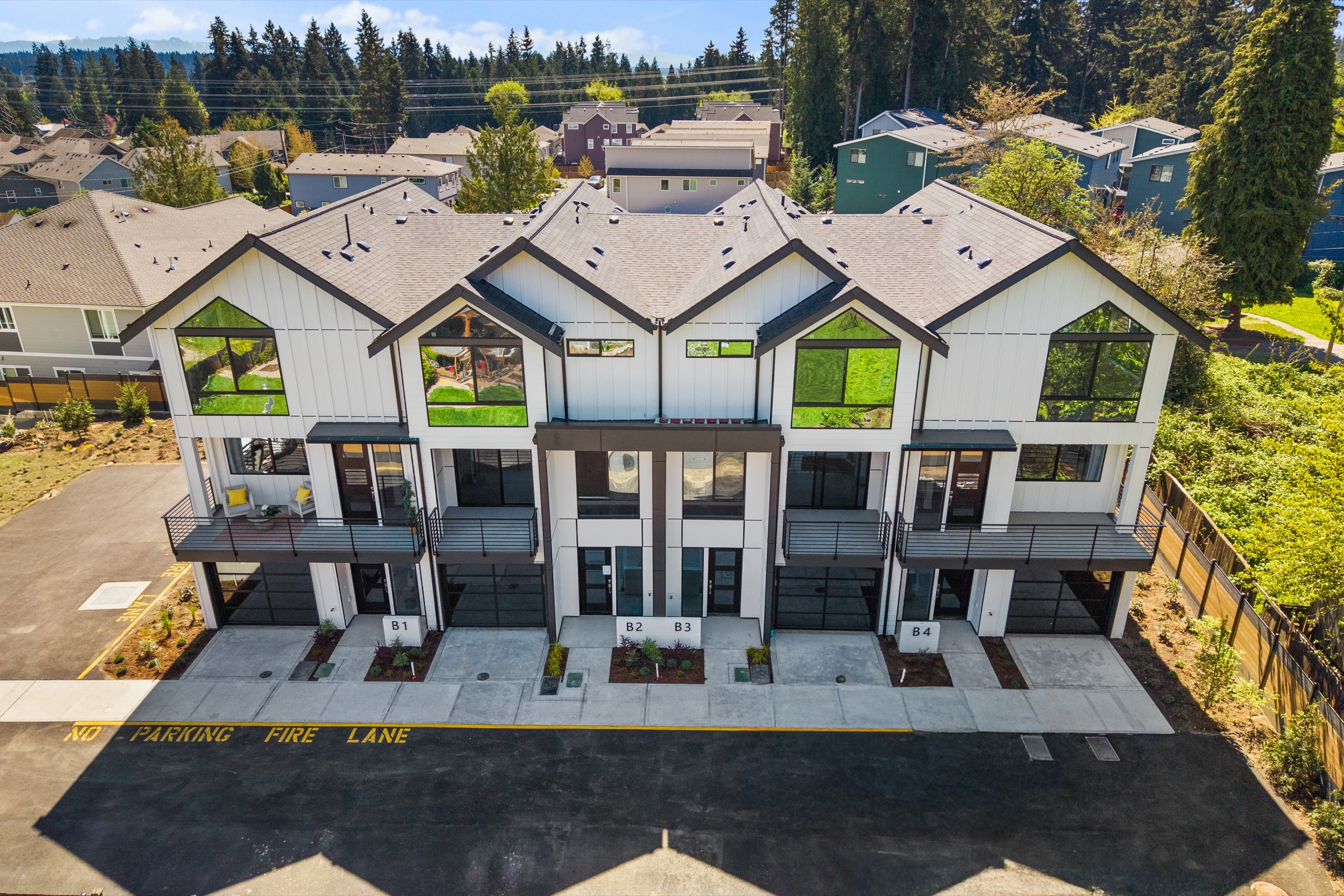 Bird's-eye view of a row of modern luxury townhomes with matching grey rooflines, individual balconies, large windows, and a shared private driveway surrounded by tall trees