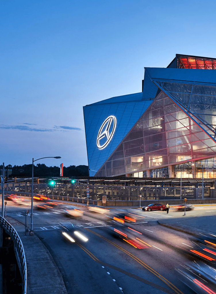 A modern stadium with glass walls and a large illuminated Mercedes-Benz logo overlooks a busy road at dusk, with light trails from passing cars and a clear evening sky.