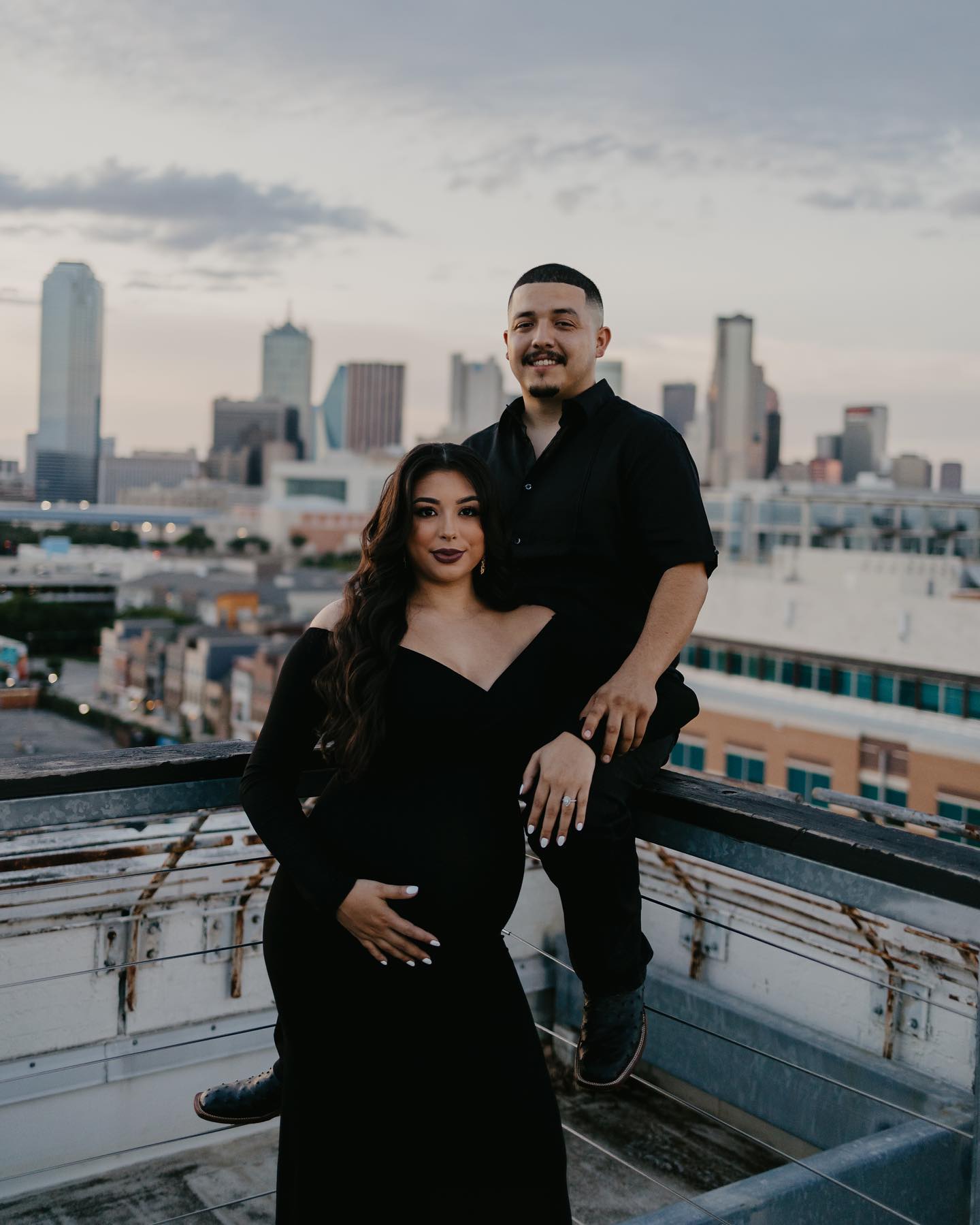 A couple stands on a rooftop with a city skyline backdrop during dusk. The woman, dressed in a black gown, cradles her baby bump, while the man in black attire stands beside her, portraying a maternity photoshoot that celebrates the anticipation of a new chapter against an urban landscape.