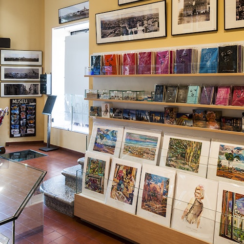 一家博物馆礼品店,墙上挂着裱好的艺术品,书架上陈列着书籍、明信片和艺术印刷品。
