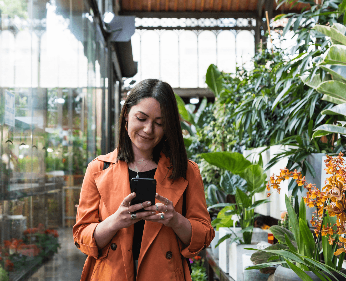 Mulher mexendo em um celular ao lado de plantas sobre prateleiras