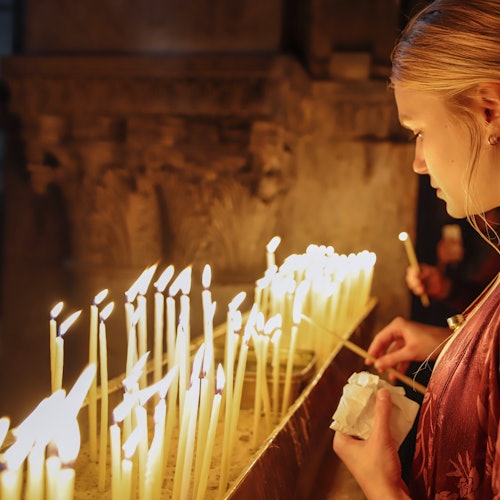 A woman lights a candle at a church altar, surrounded by many glowing candles. She holds a tissue in her hand.