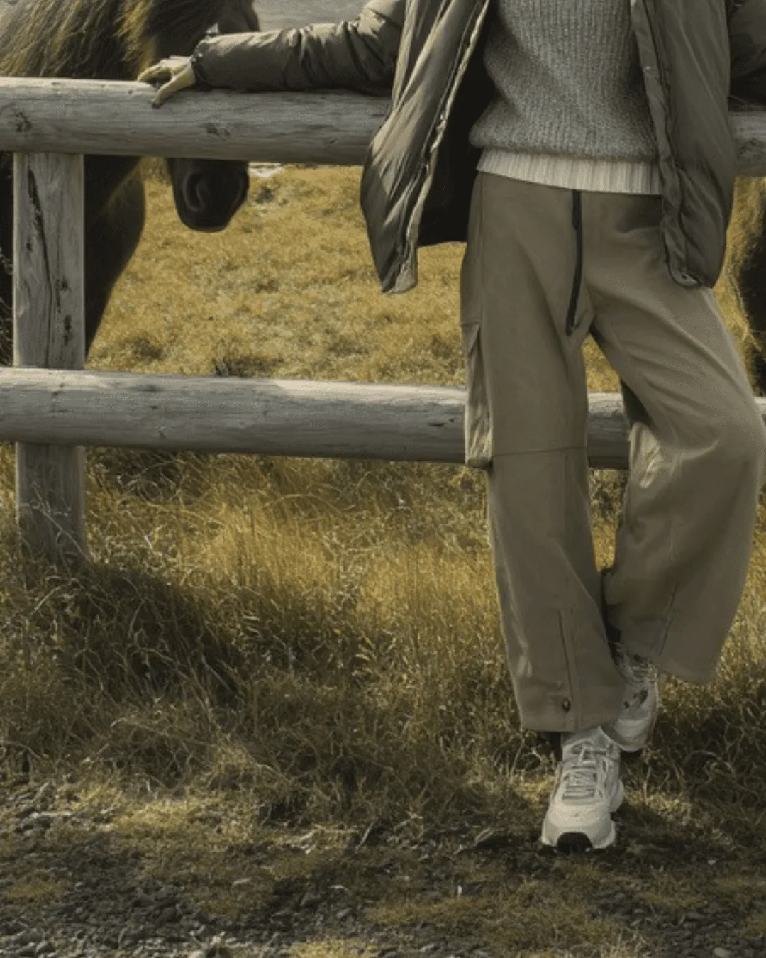 A vertical photo of a person in khaki cargo pants, an olive jacket, and white sneakers, leaning on a rustic wooden fence in a field with a horse.