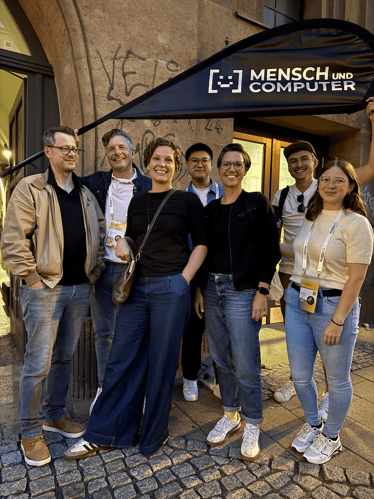 Gruppe von sieben Personen steht vor einem Gebäude mit einem schwarzen Banner mit der Aufschrift 'MENSCH UND COMPUTER'