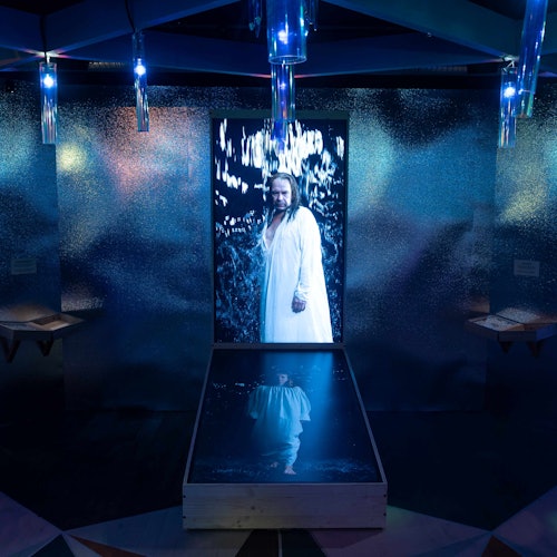 一個展示,內有穿著白袍的人的倒影,置身於一個閃亮的金屬房間內,四周環繞著藍色燈光。