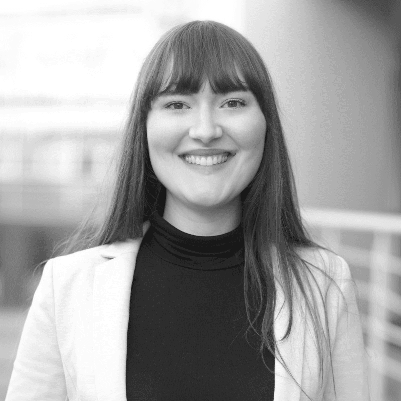 A person with curly hair and glasses smiles at the camera, seated in a cozy workspace with warm lighting.