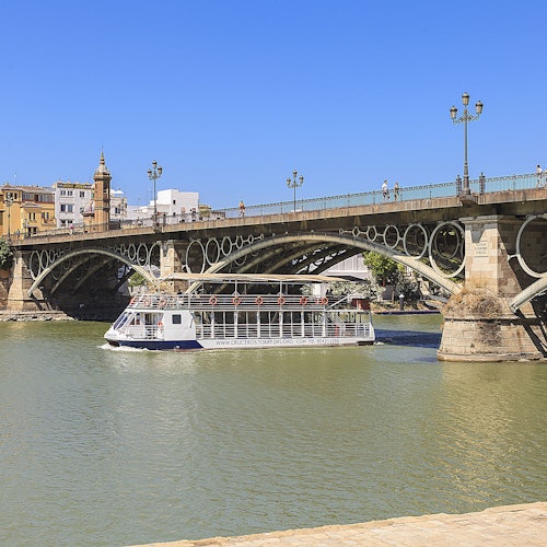 ஒரு சுற்றுலா படகு ஒரு அழகான வளைந்த பாலத்திற்கீழே பயணிக்கிறது, பின்னணியில் வரலாற்று கட்டிடங்களுடன் ஒரு உதய நாள்.