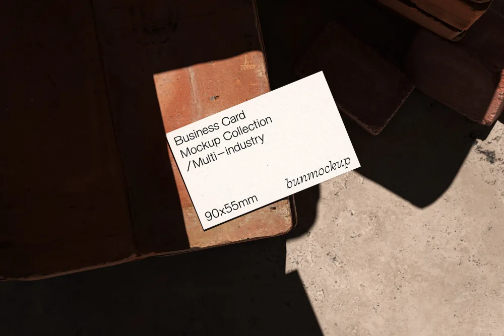Minimalist business card mockup placed on a rustic brick surface under soft, angled sunlight, creating dramatic shadows. The neutral-toned card contrasts against the earthy textures of the backdrop.