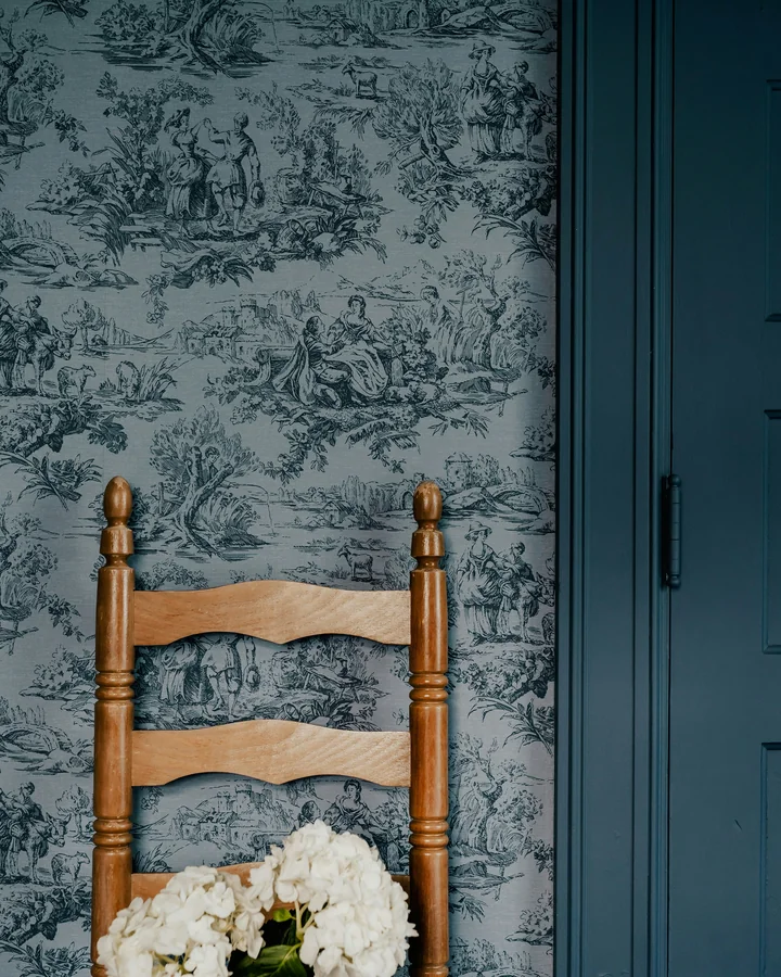 Wooden ladder-back chair with a white hydrangea arrangement against blue toile wallpaper and a dark teal door.