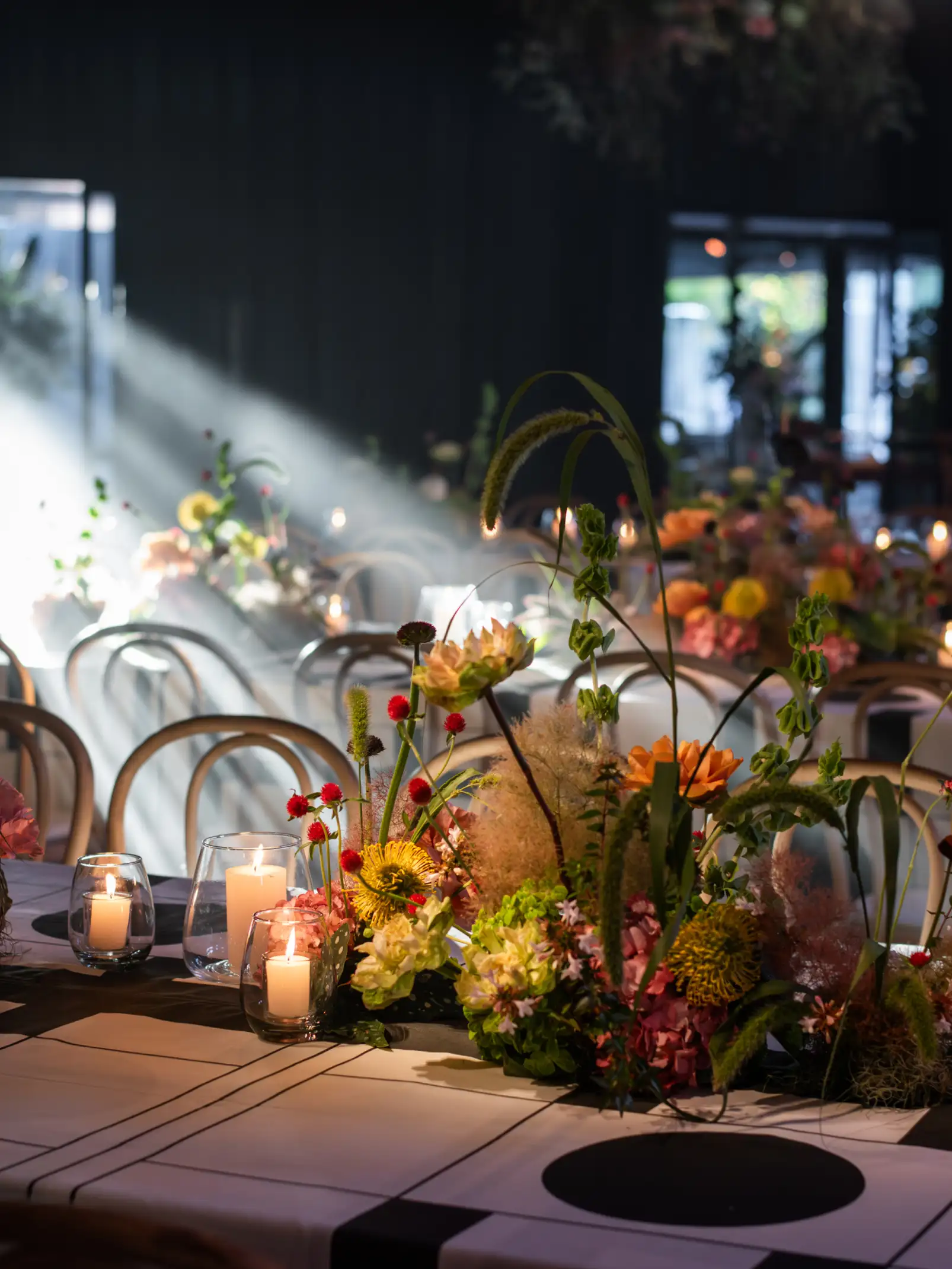 Arreglo floral contemporáneo con flores coloridas y velas encendidas en una mesa iluminada por haces de luz.