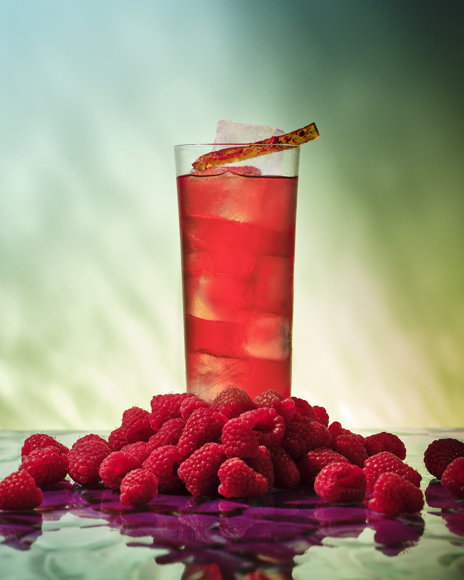 A tall glass of red drink sits on a bed of fresh raspberries, with a colorful and soft background.