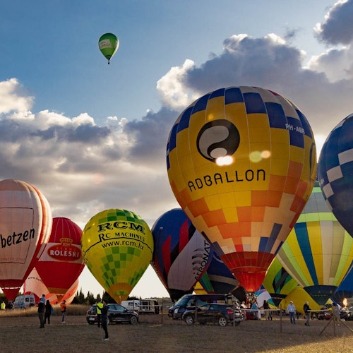 Several colorful hot air balloons on the ground with one flying in the sky under a partly cloudy sky. People and cars nearby.