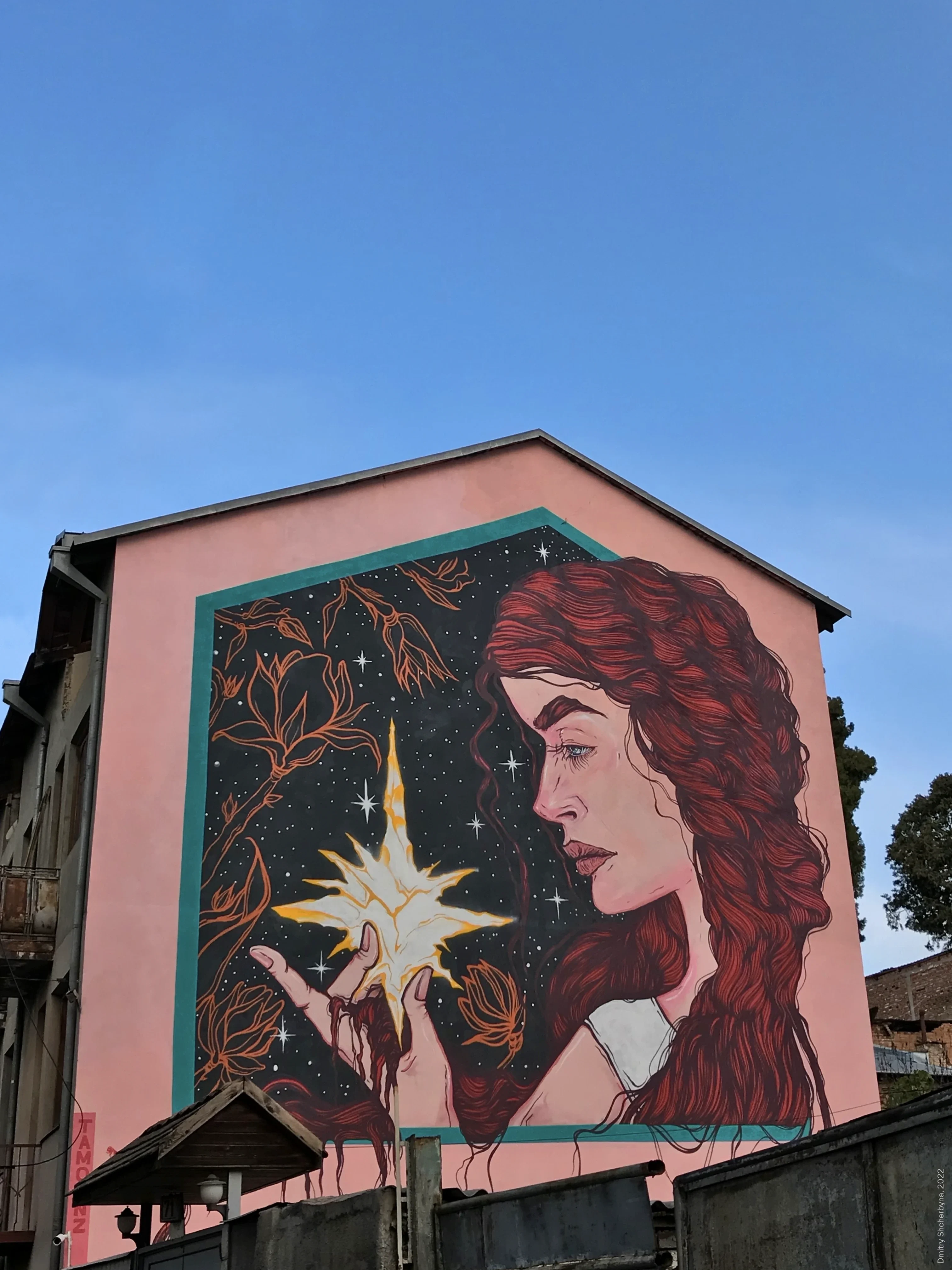 Mural on a pink building wall depicting a woman in profile, with long, flowing red curly hair holding a glowing star in her hands, against a dark night sky filled with smaller stars and stylized floral branches.