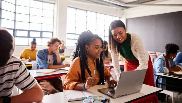 High school students receiving mentorship in STEM and RISE Research summer programs; teacher guides student typing on laptop in a bright, engaged classroom setting.