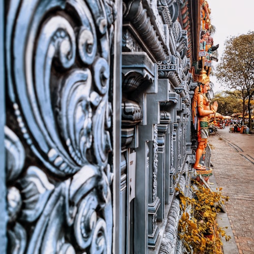 近观一座雕刻精美的寺庙墙壁，背景中是一条路径，两旁排列着色彩缤纷的雕像和树木。