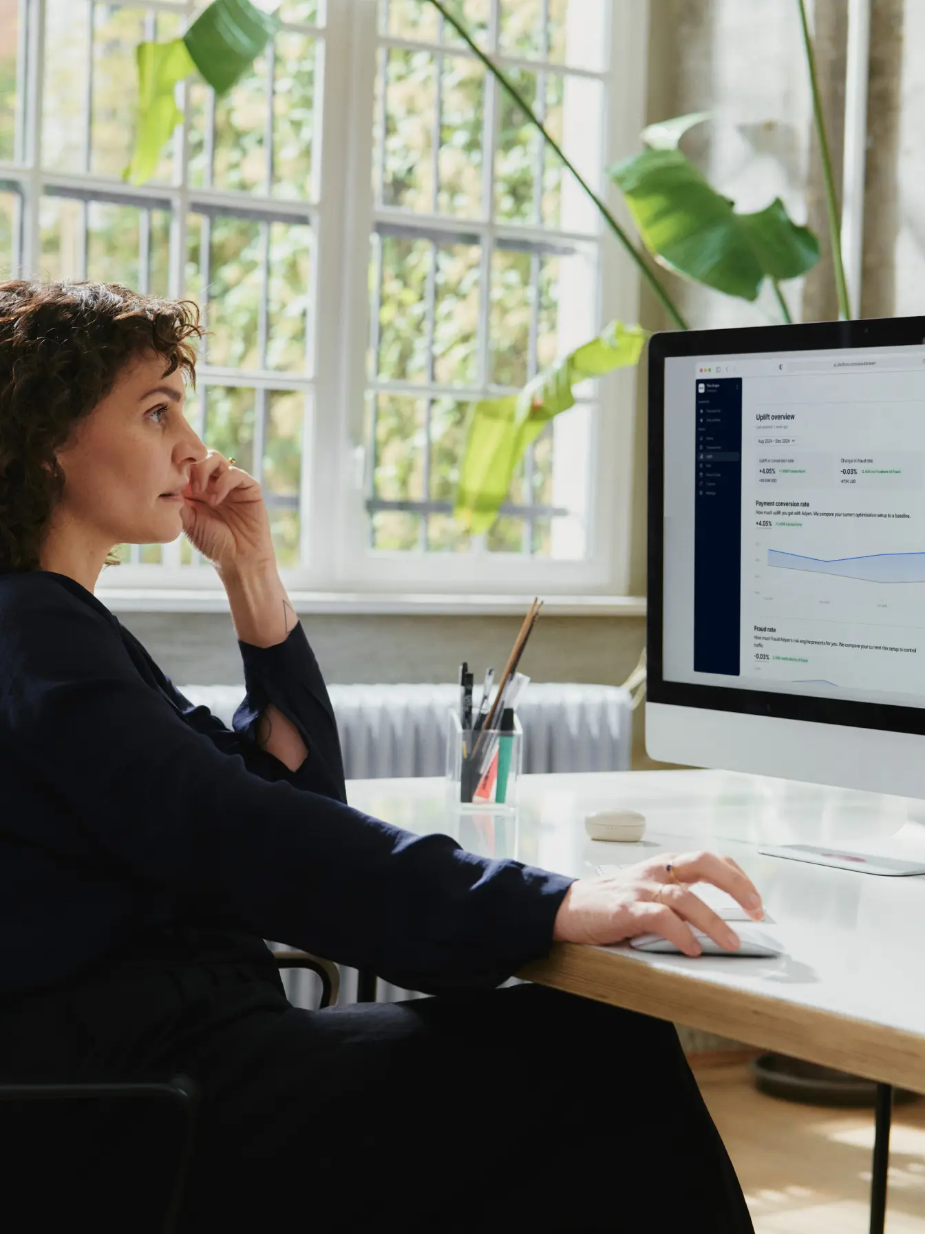 A young person with curly hair smiles at the camera while sitting in front of a laptop against a dark background.
