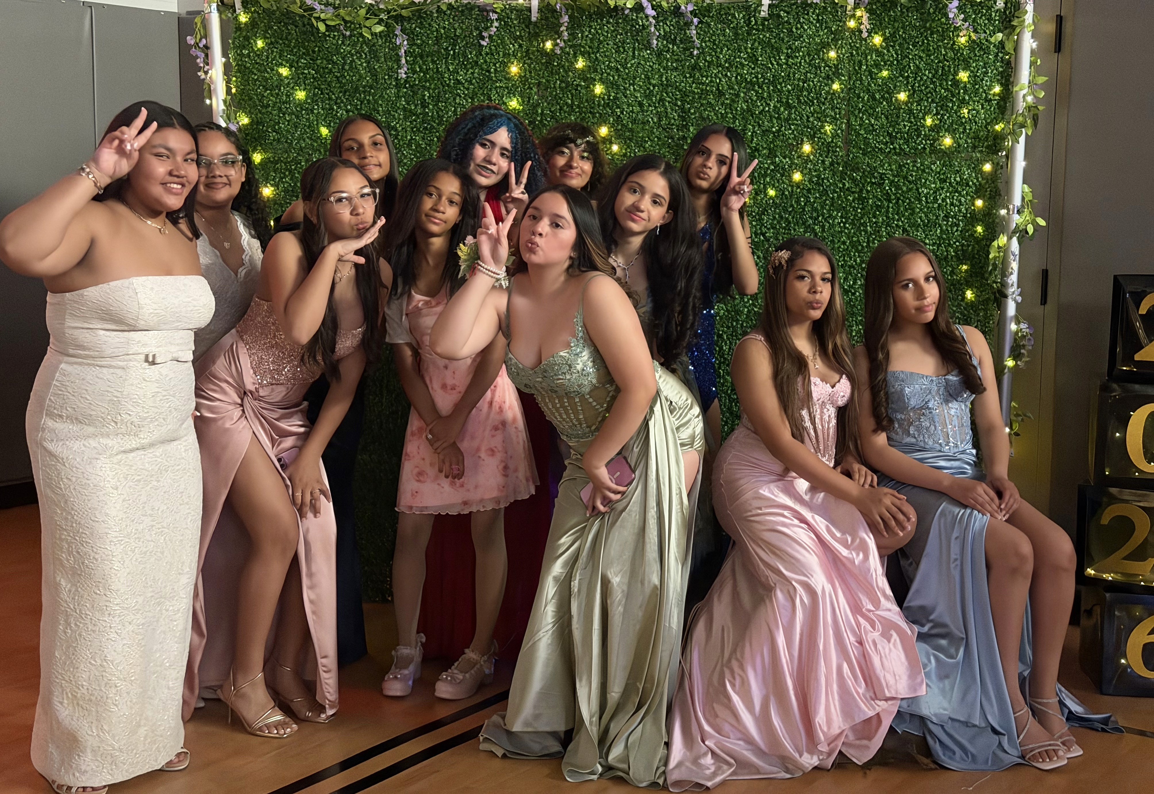 Teens pose in front of a faux grass and fairy light backdrop in formal dresses.