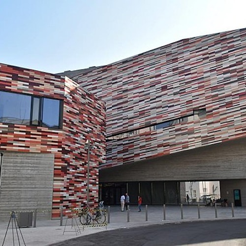 Edificio moderno con una facciata colorata e decorata, ampie finestre e un ingresso aperto. Sono visibili tre persone e diverse biciclette.