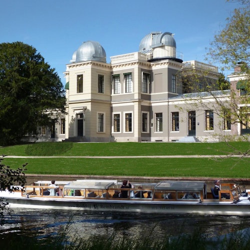 一艘載著乘客的船在平靜的河面上航行,前方是一座歷史悠久的建築,兩個圓頂的瞭望塔在晴朗的藍天下矗立。