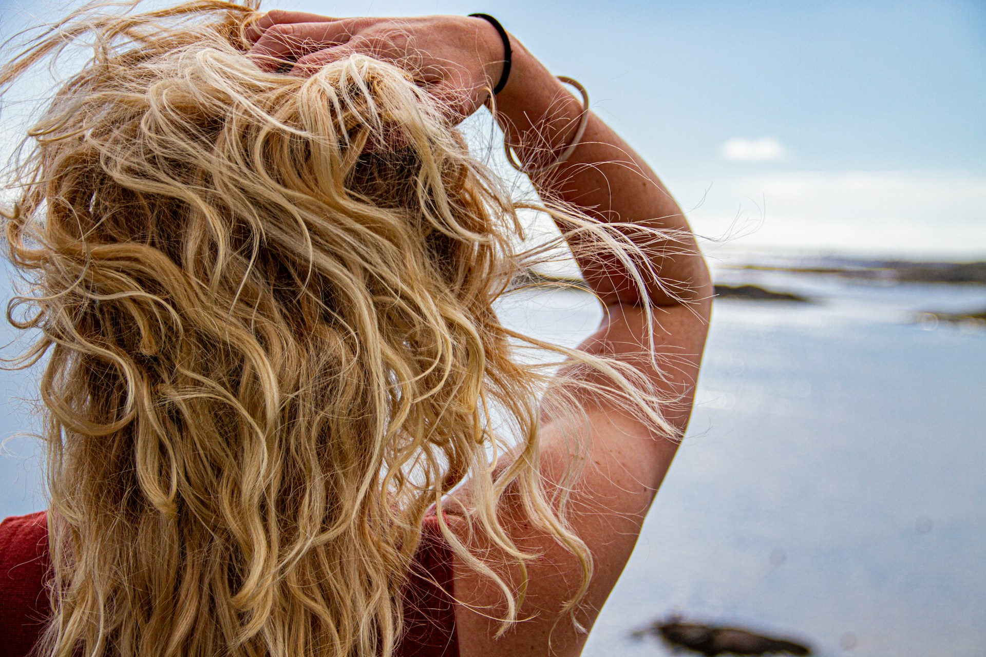 The back of a person with long, wavy blonde hair standing by the ocean with their hand in their hair.