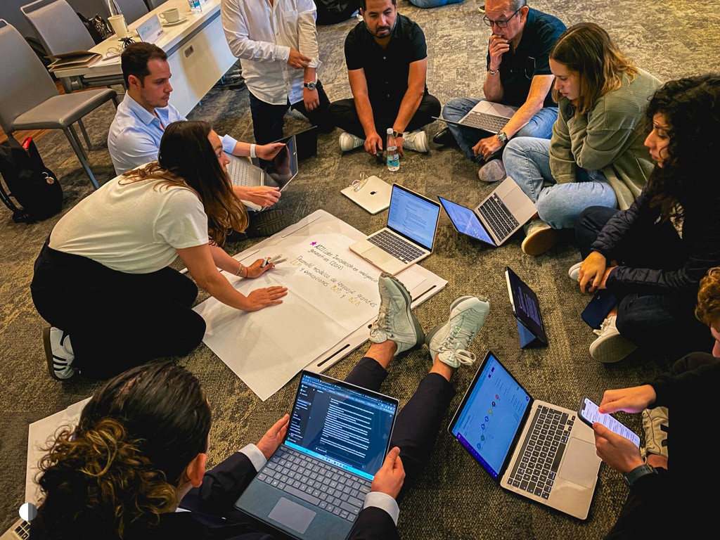 Three professional women collaborating in a bright meeting room, two using laptops and one gesturing during a discussion