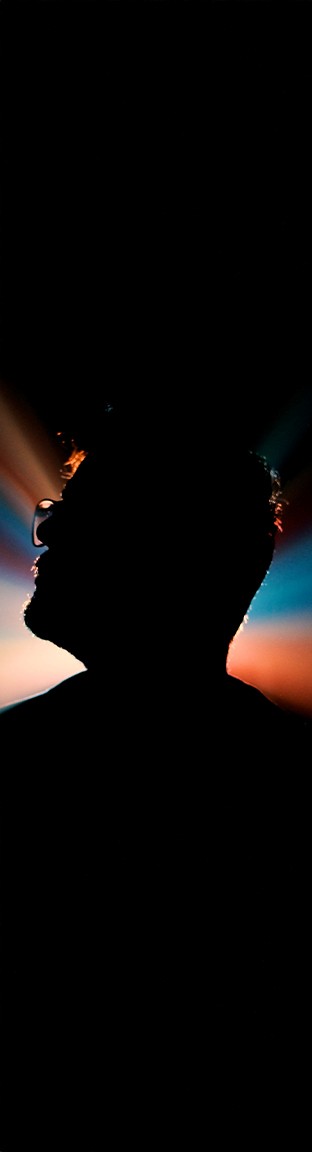 A young person with brown hair, wearing reflective silver sports sunglasses and a black sweater, looks at the camera while resting their chin on their hand.