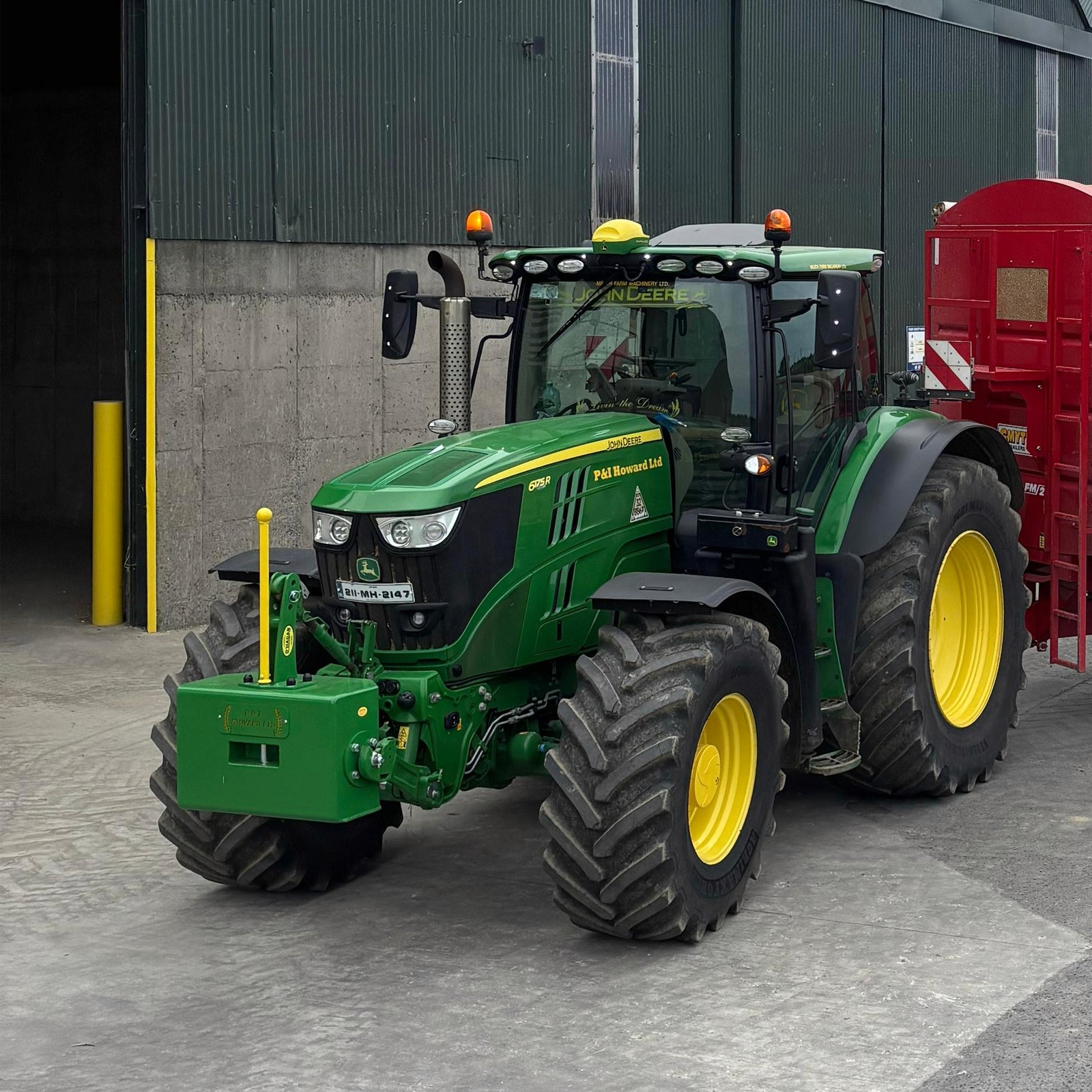 Green and yellow weight block attached to a Green John Deere with a red trailer in a yard in front of a shed