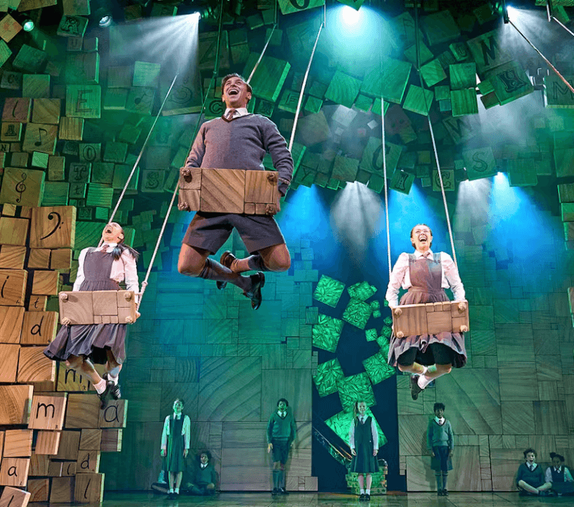 Book tickets for Matilda the Musical. Students suspended on wooden swings on a stage bathed in green lights decorated with wooden blocks that have letters, notes, punctuation marks on them.