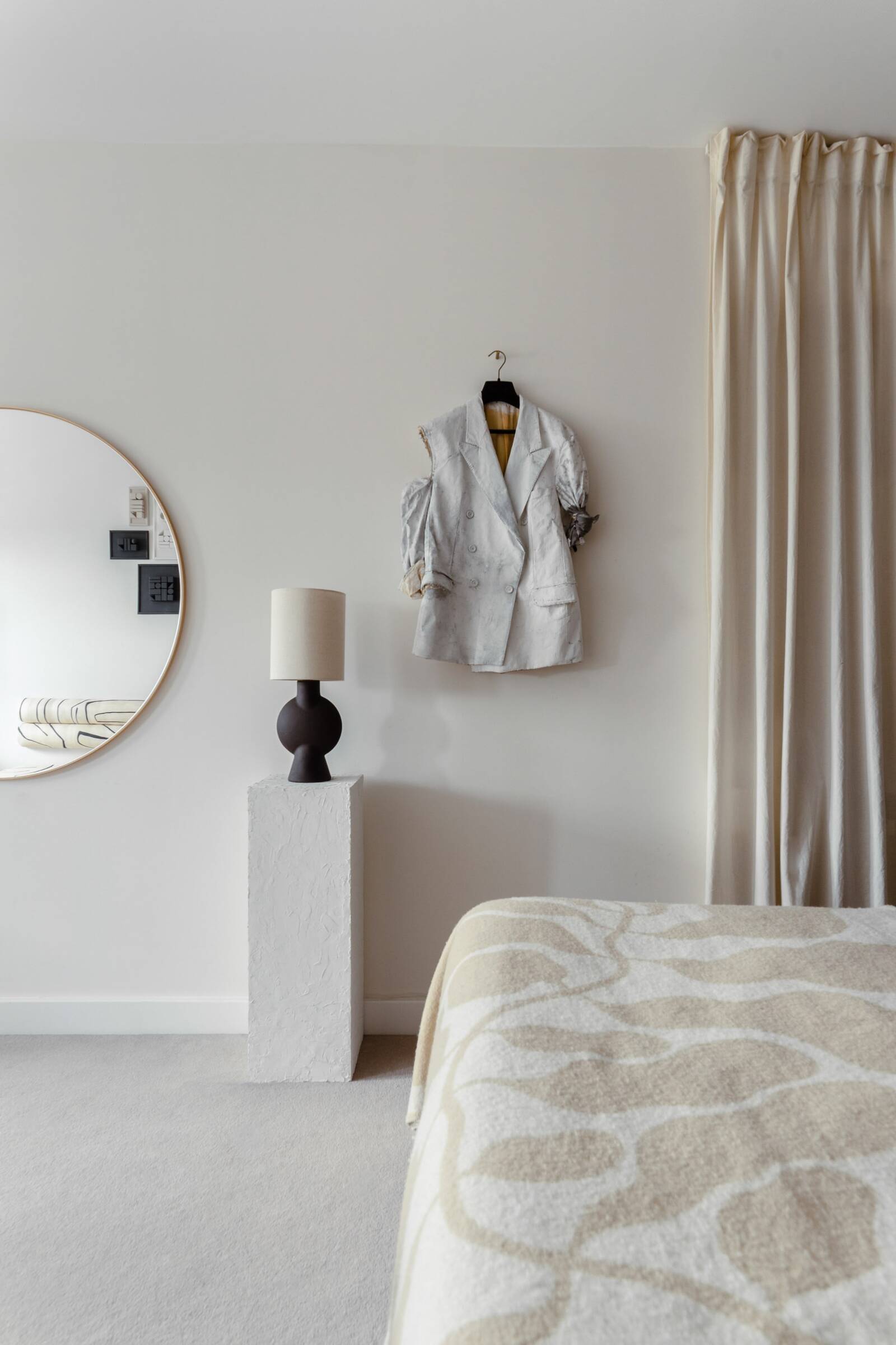 A bedroom corner with a mirror, a modern table lamp on a textured white pedestal, and a white double-breasted blazer hanging on the wall. Part of a bed with a patterned beige and white bedspread is visible.