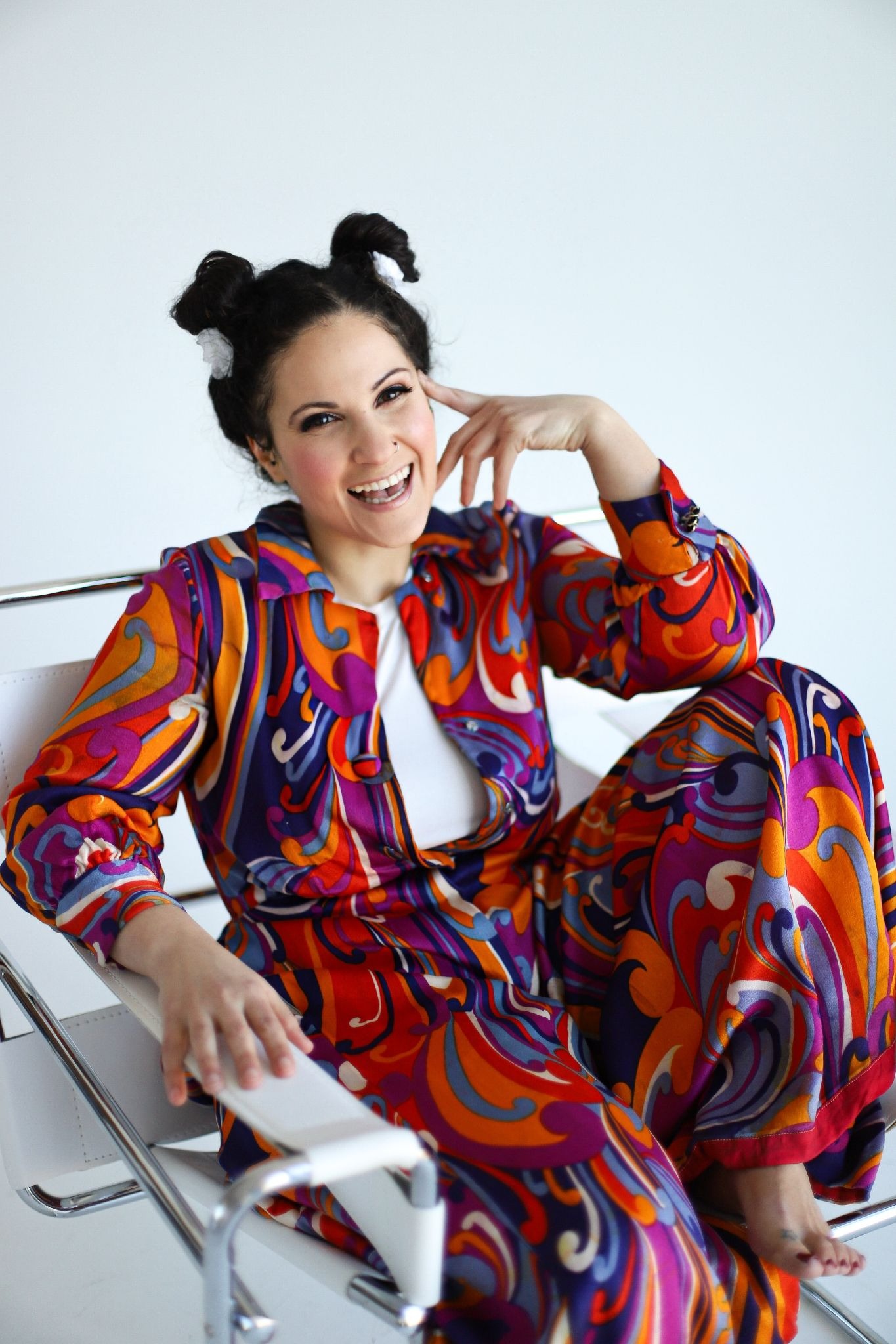 A woman smiles brightly, seated on a white chair, wearing a colorful, retro-patterned outfit in orange, purple, and red, with playful hair buns.