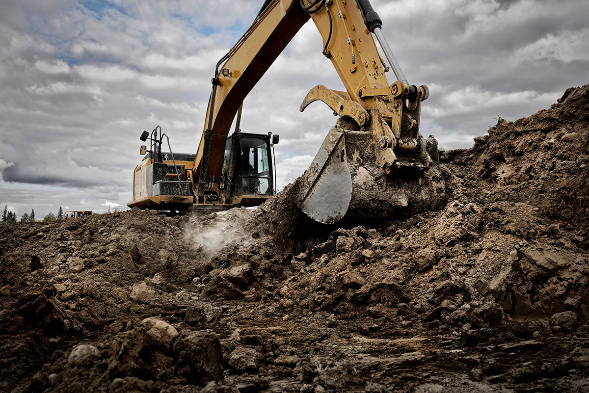 A yellow excavator digging into a large pile of dirt and rocks at a construction site under a cloudy sky.
