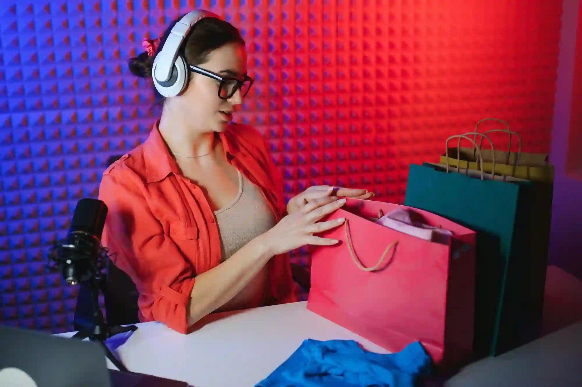 Blogger with headphones and mic unboxing items from shopping bags in a room with blue and red lighting.