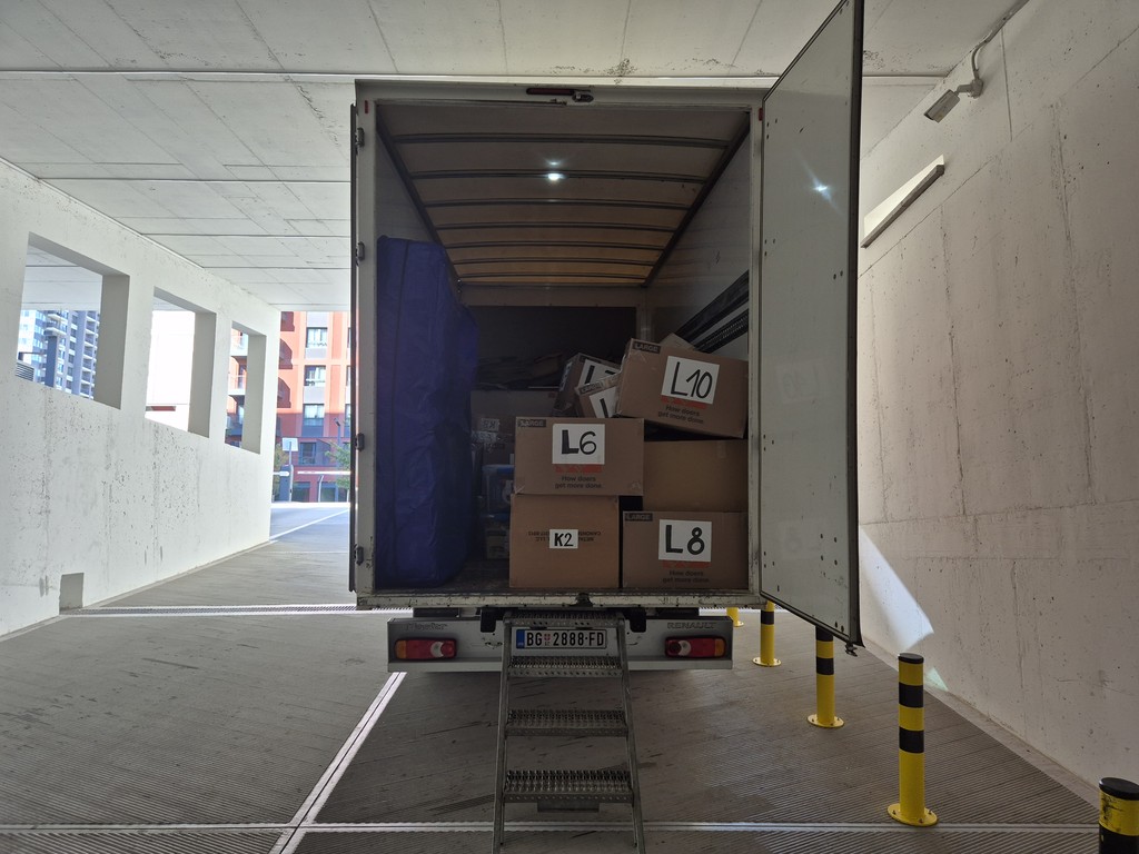 Otvoren kamion za selidbe parkiran u garaži, uredno složene kutije i zaštitni materijal unutra, spremni za istovar u moderno stambeno naselje — profesionalna usluga selidbi u Beogradu.