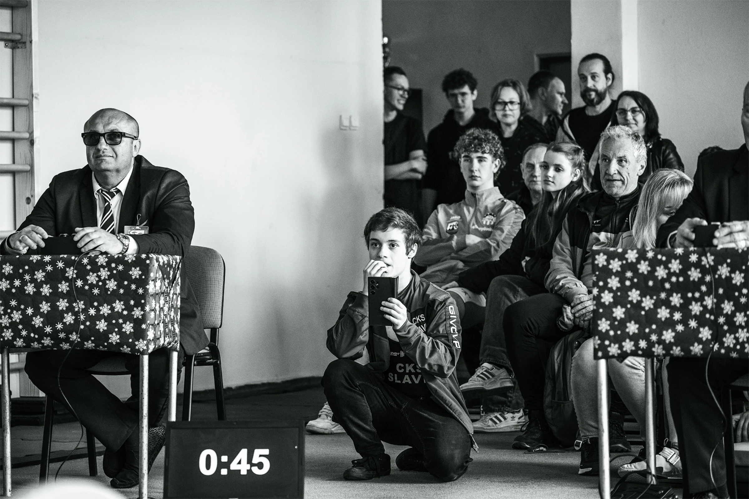 Czarno-białe zdjęcie z zawodów: sędzia w okularach przeciwsłonecznych przy stole i młoda osoba filmująca telefonem.