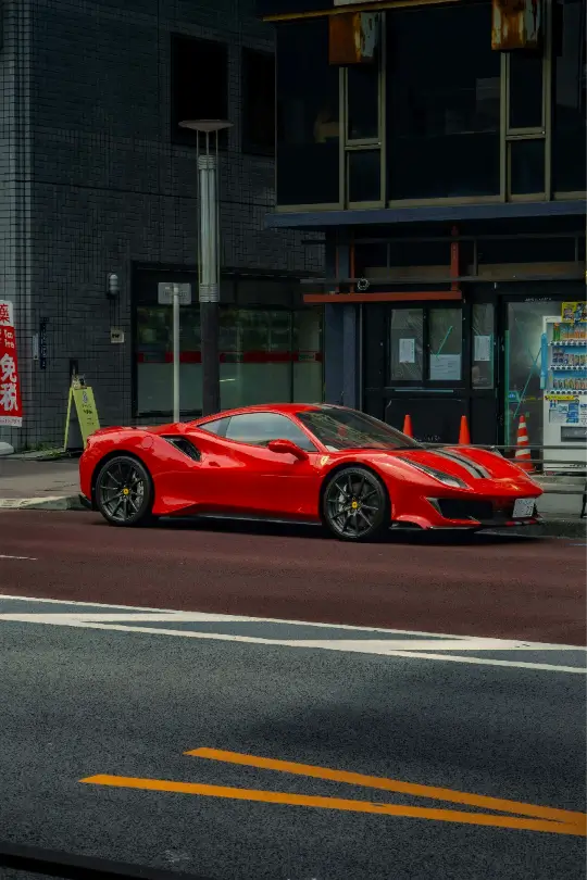 A red Ferrari 488 Pista parked on a city street in Japan, featuring black racing stripes and dark alloy wheels against a backdrop of urban buildings.