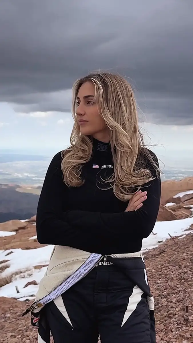 Emelia Hartford in her black racing outfit, confidently standing atop a snowy mountain peak.