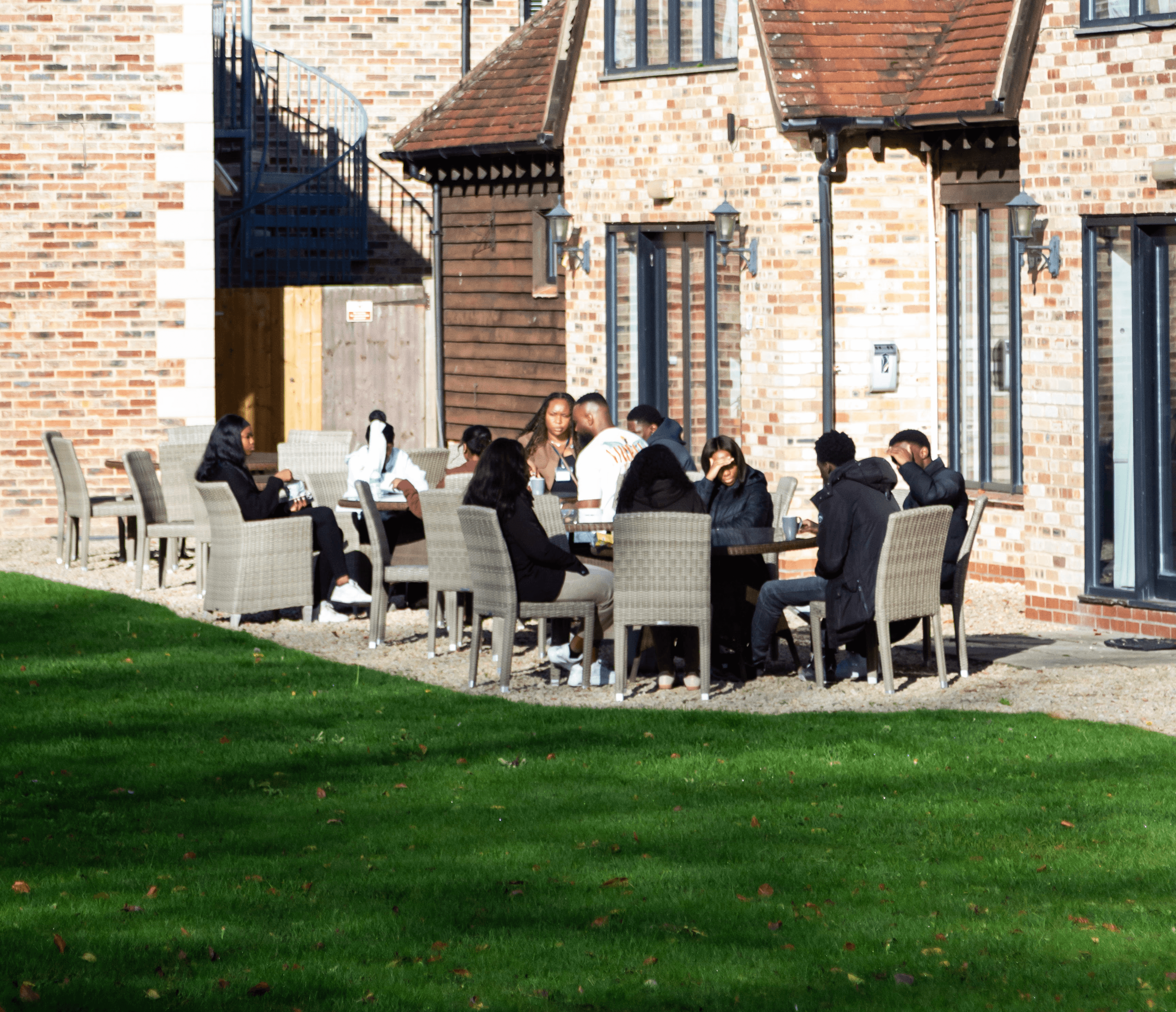 A group of people seated around a table outside a building, with a green lawn in the foreground.